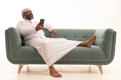 Making a video call via mobile, communicating with family and friends, browsing social media, using a modern technological device, a portrait of a Saudi Arabian Gulf man wearing a white thobe and a headscarf sitting on a sofa working on a mobile phone, white background.
