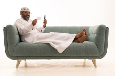 Online shopping, the concept of remote payment and settlement via mobile applications, the use of electronic bank cards, a portrait of a Saudi Gulf Arab man wearing a white thobe and a headscarf sitting on a sofa holding a mobile phone, white background.