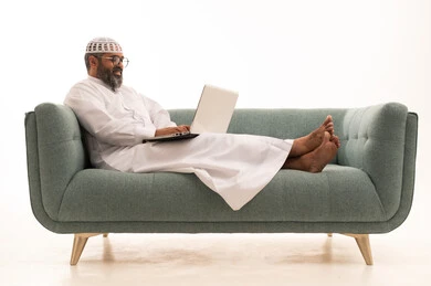 Browsing applications and electronic programs via the laptop, using modern and advanced technological devices, managing business remotely, a portrait of a Saudi Arabian Gulf man wearing a white thobe and a headscarf sitting on the sofa working on a laptop, white background.