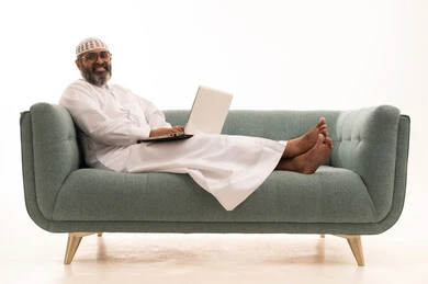 Remote business management, browsing applications and electronic programs via laptop, using modern and advanced technological devices, a portrait of an Arab Gulf Saudi man wearing a white thobe and a headscarf sitting on a sofa working on a laptop, white background.