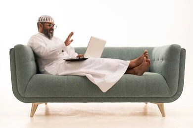 Making a video call, raising the palm with expressions of peace and greeting, the development of technology and technical devices, a portrait of an Arab Gulf Saudi man wearing a white thobe and a headscarf sitting on a sofa working on a laptop, white background.