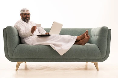 The concept of remote payment and settlement through laptop applications, the use of electronic bank cards, the evolution of technology and technical devices, a portrait of an Arab Gulf Saudi man wearing a white thobe and a headscarf sitting on a sofa working on a laptop, white background.