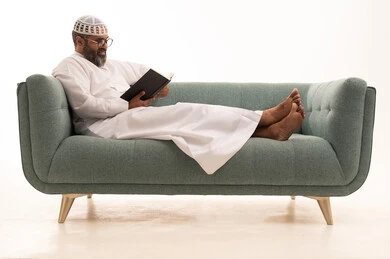 The hobby of reading and exploration, spending free time learning and cultural activities, the concept of education and scientific research, a portrait of an Arab Gulf Saudi man wearing a white thobe and a head cap sitting on a sofa holding a book, gestures of engagement and focus, white background.