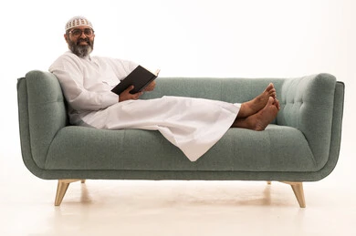 النظر الى الكاميرا بتعابيرالسرور، هواية المطالعة والاستكشاف، قضاء وقت الفراغ بالتعلم والثقافة، مفهوم التثقيف والبحث العلمي،  بورتريه لرجل عربي خليجي سعودي يرتدي الثوب الابيض و الطاقية يجلس على الاريكة يمسك كتاب، خلفية بيضاء