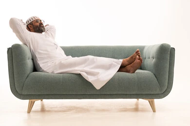 إيماءات الراحة والإسترخاء، الشعور بالتعب و الخمول والحاجة إلى النوم, بورتريه لرجل عربي خليجي سعودي يرتدي الثوب الابيض و الطاقية يجلس على الاريكة يضع يدية وراء رأسه بتعابير التأمل والسرور، النظر للأعلى إلى شيء ما، خلفية بيضاء