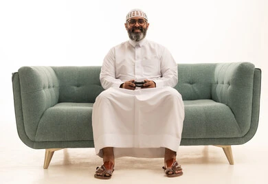 الاندماج والتفاعل مع ألعاب البلايستيشن، بورتريه لرجل عربي خليجي سعودي يرتدي الثوب الابيض و الطاقية يجلس على الاريكة يحمل بيده عصا التحكم يقوم باللعب في ألعاب الفيديو، اجهزة وتقنيات الكترونية حديثة ترفيهيه, قضاء وقت ممتع ومسلي باللعب، خلفية بيضاء