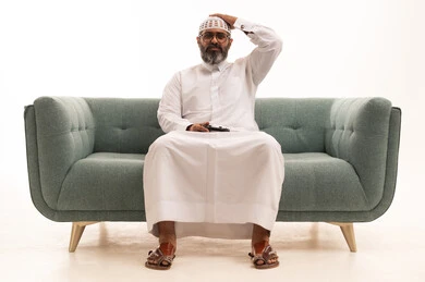 ايماءات الخسارة والهزيمة ،استخدام الأجهزة التكنولوجية والتقنيات الحديثة، الاستمتاع باللعب بالعاب الفيديو الترفيهية، بورتريه لرجل عربي خليجي سعودي يرتدي الثوب الابيض و الطاقية يجلس على الاريكة ويمسك بيده عصا التحكم بتعابير الحزن، خلفية بيضاء