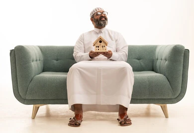 Looking at something, the pursuit of owning a new house, buying and selling real estate, the pursuit of owning a new house, strong family ties, a portrait of a Saudi Arabian Gulf man wearing a white thobe and a head cap sitting on a sofa holding a small wooden model of a house with gestures of joy and hope, white background.