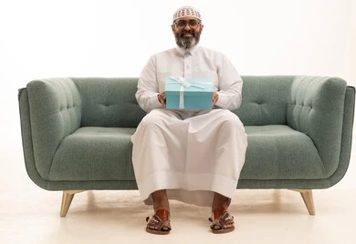 تقديم الهدايا في العيد والمناسبة الخاصة، التعبير عن الحب والتقدير والاحترام، بورتريه لرجل عربي خليجي سعودي يرتدي الثوب الابيض و الطاقية يجلس على الاريكة يحمل صندوق الهدية ينظر  بإيماءات السرور، مفهوم الشكر والامتنان، خلفية بيضاء