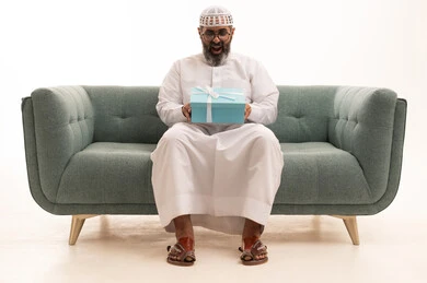 التعبير عن الحب والتقدير والاحترام، بورتريه لرجل عربي خليجي سعودي يرتدي الثوب الابيض و الطاقية يجلس على الاريكة يحمل صندوق الهدية ينظر  بإيماءات السرور، تقديم الهدايا في العيد والمناسبة الخاصة، مفهوم الشكر والامتنان، خلفية بيضاء