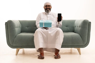 A blank black screen displayed on the mobile, expressing love, appreciation, and respect, a portrait of a Gulf Arab Saudi man wearing a white thobe and a headscarf sitting on the sofa holding a gift box looking with gestures of joy, the concept of gratitude and thankfulness, white background.