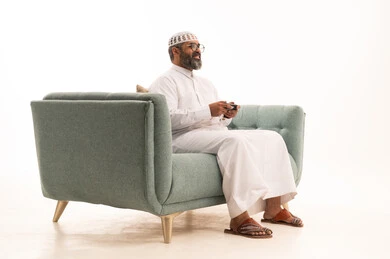 Integration and interaction with PlayStation games, a portrait of a Gulf Arab Saudi man wearing a white thobe, a cap, and a scarf sitting on a sofa holding a game controller playing video games, modern entertainment electronic devices and technologies, spending enjoyable and entertaining time playing, white background.