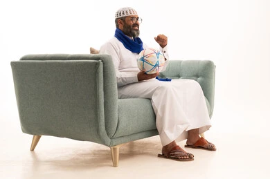 تعابير وجه ويدين تدل على الحماس والتشجيع، الشغف بمتابعة فريق كرة القدم، الانتصار وتحقيق الأهداف وفوز الفريق المفضل بالبطولة، بورتريه لرجل عربي خليجي سعودي يرتدي الثوب الابيض و الطاقية والوشاح يجلس على الاريكة يمسك كره القدم، الجسم بالطول الكامل، خلفية بيضاء