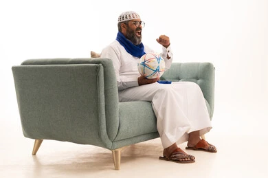 The passion for following a football team, victory, achieving goals, and the favorite team's win in the championship, a portrait of an Arab Gulf Saudi man wearing a white thobe, a cap, and a scarf sitting on a sofa holding a football, facial and hand expressions indicating enthusiasm and encouragement, full-body, white background.