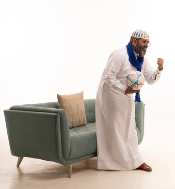 الانتصار وتحقيق الأهداف وفوز الفريق المفضل بالبطولة، الشغف بمتابعة فريق كرة القدم، بورتريه لرجل عربي خليجي سعودي يرتدي الثوب الابيض و الطاقية والوشاح يقف بجانب الاريكة يمسك كره القدم، تعابير وجه ويدين تدل على الحماس والتشجيع، الجسم بالطول الكامل، خلفية بيضاء