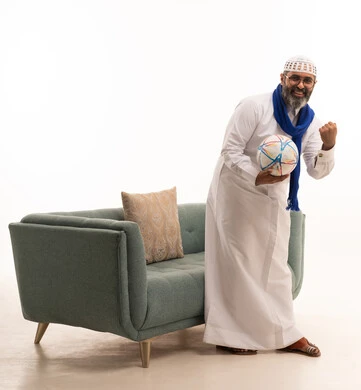 Integration through encouraging football, gestures of victory and triumph, a portrait of a Saudi Gulf Arab man wearing a white thobe, a headscarf, and a shawl standing next to a sofa holding a football, raising his hands with interaction and enthusiasm, a full-body image, white background.