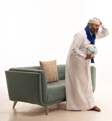 إيماءات الخسارة والفشل، مشاهدة ومتابعة مباراة كرة القدم، بورتريه لرجل عربي خليجي سعودي يرتدي الثوب الابيض و الطاقية والوشاح  يقف بجانب الاريكة يمسك كره القدم ويضع يده على رأسه بتعابير الحزن والتفاعل مع المنتخب، الجسم بالطول الكامل، خلفية بيضاء