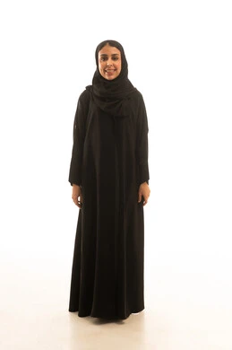 A full-length portrait with a white background of a Gulf Arab woman wearing a black abaya, standing and smiling in front of the camera.