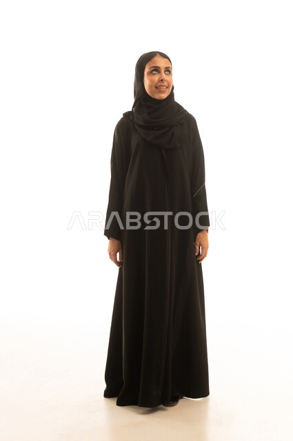 A Saudi Arabian Gulf woman wearing a black abaya and a hijab is looking at something, full-body portrait with a white background.