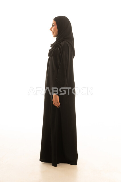 A Saudi Arabian Gulf woman wearing a black abaya, standing sideways in front of the camera, full-body portrait with a white background, showing a gesture of happiness and a smile.