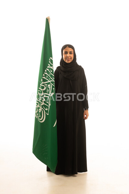 A Saudi Arabian Gulf woman wearing a black abaya and a hijab is holding the green flag of Tawheed, the Saudi flag. It is a full-body portrait with a white background, looking directly at the camera.