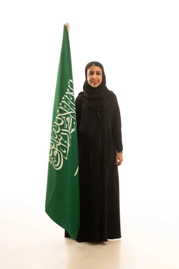 A Saudi Arabian Gulf woman wearing a black abaya and a hijab is holding the green flag of Tawheed, the Saudi flag. It is a full-body portrait with a white background, looking directly at the camera.