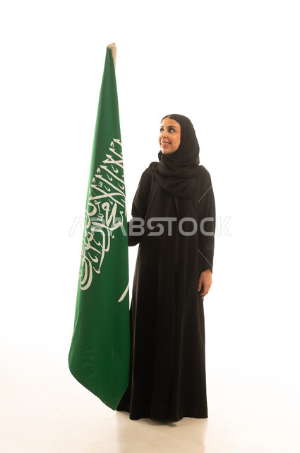 راية العزة والتوحيد الراية الخضراء ,امرأة عربية خليجية سعودية محجبة ترتدي عباءة سوداء تقف بجانب العلم السعودي ,بورتريه خلفية بيبضاء الجسم بالطول الكامل ,النظر الى شئ ما