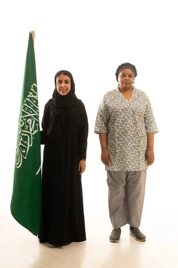 A Saudi Arabian Gulf woman wearing a black abaya and holding the Saudi flag, a portrait with a white background of an African domestic worker with dark skin wearing a gray uniform, full body, looking at the camera.