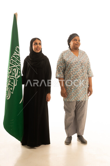 عاملة منزلية افريقية سمراء البشرة ترتدي يونيفورم باللون الرمادي تقف بجانب امراة عربية خليجية سعودية محجبة ترتدي عباءة سوداء تمسك العلم السعودي ,بورتريه خلفية بيضاء الجسم بالطول الكامل ,النظر الى شئ ما