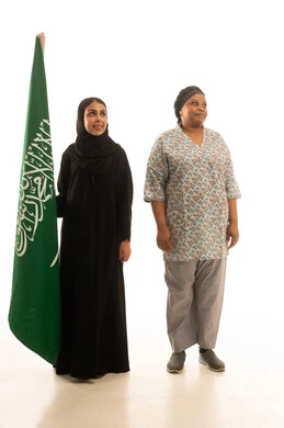 عاملة منزلية افريقية سمراء البشرة ترتدي يونيفورم باللون الرمادي تقف بجانب امراة عربية خليجية سعودية محجبة ترتدي عباءة سوداء تمسك العلم السعودي ,بورتريه خلفية بيضاء الجسم بالطول الكامل ,النظر الى شئ ما