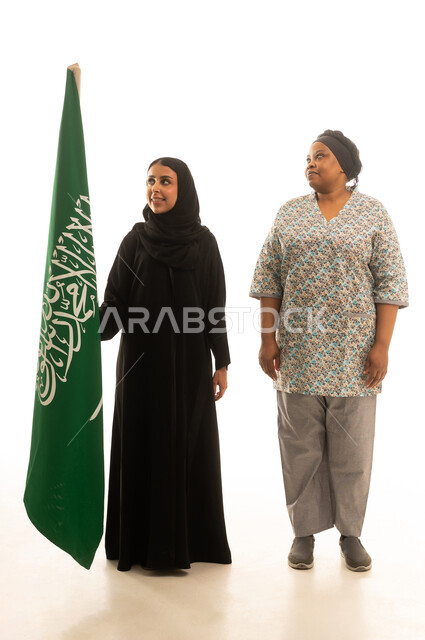 الوقوف بشموخ بجانب راية التوحيد ,امراة عربية خليجية سعودية محجبة ترتدي عباءة سوداء تمسك العلم السعودي مع العاملة المنزلية الافريقية سمراء البشرة ترتدي يونيفورم باللون الرمادي , بورتريه خلفية بيضاء الجسم بالطول الكامل