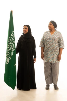 الوقوف بشموخ بجانب راية التوحيد ,امراة عربية خليجية سعودية محجبة ترتدي عباءة سوداء تمسك العلم السعودي مع العاملة المنزلية الافريقية سمراء البشرة ترتدي يونيفورم باللون الرمادي , بورتريه خلفية بيضاء الجسم بالطول الكامل