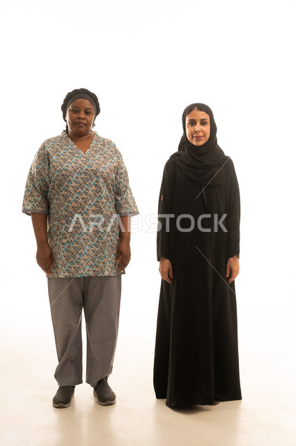 الوقوف جنبًا الى جنب والنظر للكاميرا ,امرأة عربية خليجية سعودية محجبة ترتدي عباءة سوداء ,عاملة منزلية افريقية سمراء البشرة ترتدي يونيفورم اللون الرمادي ,بورتريه خلفية بيضاء الجسم بالطول الكامل