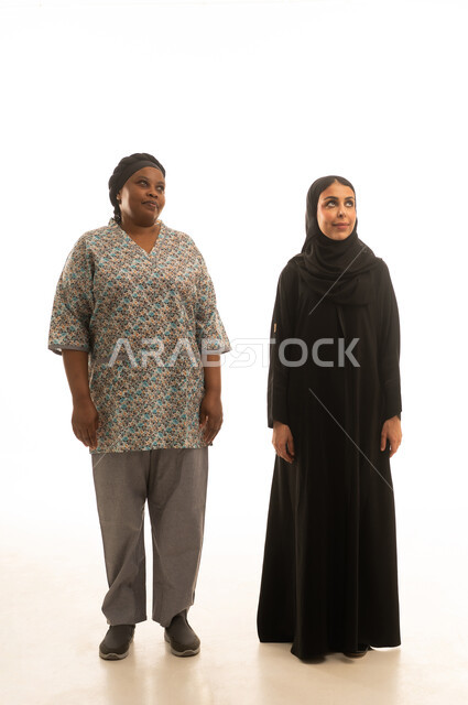 A portrait with a white background featuring a Saudi Arabian Gulf woman wearing a black abaya and a dark-skinned African housemaid in a gray uniform, full-body, looking at something.