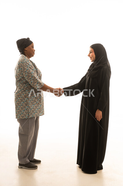 A handshake is a sign of peace. A Saudi Arabian Gulf woman wearing a black abaya greets an African domestic worker with dark skin who is wearing a gray uniform. Full-body portrait with a white background.