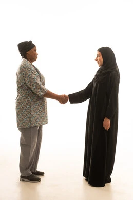 A handshake is a sign of peace. A Saudi Arabian Gulf woman wearing a black abaya greets an African domestic worker with dark skin who is wearing a gray uniform. Full-body portrait with a white background.