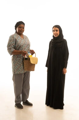 A portrait with a white background of a Saudi Arabian Gulf woman wearing a black abaya and a hijab, standing next to an African domestic worker with brown skin wearing a gray uniform, holding a wicker basket filled with household care supplies, full body.