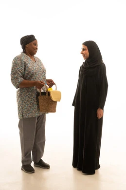 A Saudi Arabian Gulf woman wearing a black abaya and a hijab is following household chores, a full-body portrait against a white background of an African domestic worker with dark skin wearing a gray uniform, carrying a wicker basket filled with cleaning tools.