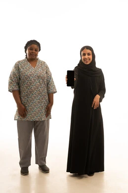 امرأة عربية خليجية سعودية محجبة ترتدي عباءة سوداء مع عرض شاشة هاتف محمول سوداء فارغة للكاميرا ,عاملة منزلية افريقية سمراء البشرة ترتدي يونيفورم باللون الرمادي ,بورتريه خلفية بيضاء الجسم بالطول الكامل , النظر للكاميرا