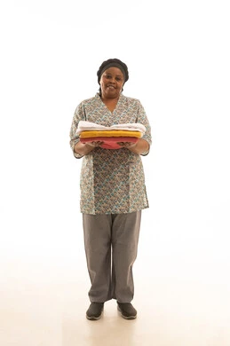 الانتهاء من الاعمال اليومية بسعادة ,بورتريه خلفية بيضاء لعاملة منزلية افريقية سمراء البشرة ترتونيفورم باللون الرمادي تحمل المناشف و الملابس النظيفة ,الجسم بالطول الكامل