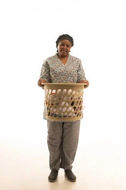 An African housekeeper with dark skin wearing a gray uniform holding a plastic laundry basket, full-body portrait against a white background, smiling and looking at the camera.