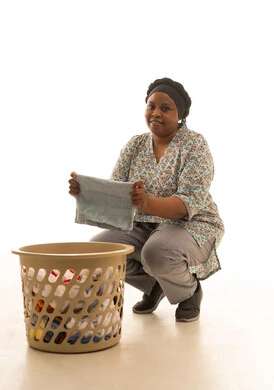 النظر للكاميرا مع الانتهاء من طي الملابس ,عاملة منزلية افريقية سمراء البشرة ترتدي يونيفورم باللون الرمادي تجلس القرفصاء امام سلة ملابس بلاستيكية ,بورترية خلفية بيضاء الجسم بالطول الكامل