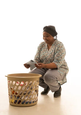 طي و ترتيب الملابس بعد الانتهاء من غسلها ,بورتريه خلفية بيضاء لعاملة منزلية افريقية سمراء البشرة ترتدي يونيفورم باللون الرمادي تجلس امام سلة الملابس البلاستيكية ,الجسم بالطول الكامل