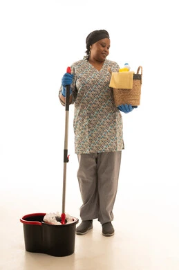 An African woman with dark skin is looking at a wicker basket containing household cleaning supplies. A colored domestic worker is wearing a gray uniform and blue leather gloves, holding a manual floor mop. A full-length portrait with a white background.