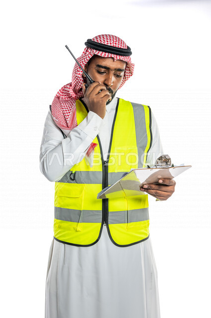 بورتريه لمهندس عربي خليجي سعودي ، يرتدي الزي السعودي التقليدي وسترة الحماية خاصة بالعمل ، يستخدم جهاز اتصال لاسلكي ، يحمل بيده تقارير ، الإشراف ومتابعة العمل ، إعطاء أوامر خاصة بالعمل ، مهن و وظائف هندسية ، ادارة المشاريع المعمارية الهندسية ، خلفية بيضاء