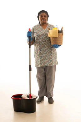 بورتريه خلفية بيضاء لعاملة منزلية افريقية سمراء البشرة ترتدي يونيفورم باللون الرمادي تحمل مستلزمات النظافة داخل سلة من القش ,استخدام ممسحة الارضية في اعمال التنظيف , الجسم بالطول الكامل