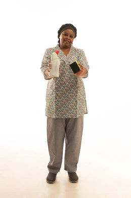 العناية و الاهتمام بالمنزل ,بورتريه خلفية بيضاء لشغالة افريقية ملونة سمراء البشرة ترتدي يونيفورم باللون الرمادي تعرض زجاجة من سائل غسيل الصحون و اسفنجة سوداء للكاميرا ,الجسم بالطول الكامل ,الابتسامة للكاميرا