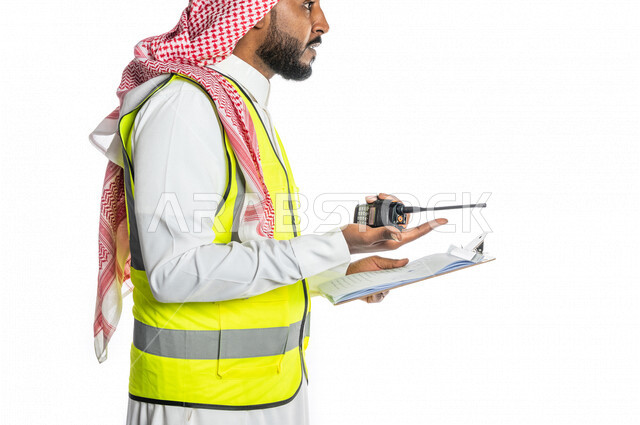 بورتريه لمهندس عربي خليجي سعودي ، يرتدي الزي السعودي التقليدي وسترة الحماية خاصة بالعمل ، يستخدم جهاز اتصال لاسلكي ، يحمل بيده تقارير ، الإشراف ومتابعة العمل ، إعطاء أوامر خاصة بالعمل ، مهن و وظائف هندسية ، ادارة المشاريع المعمارية الهندسية ، خلفية بيضاء