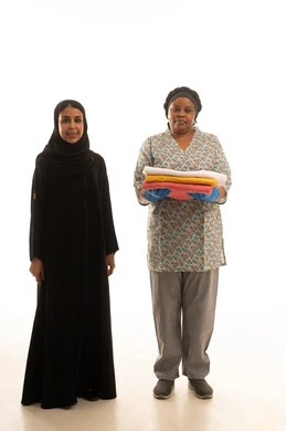 A full-length portrait on a white background of a Saudi Arabian Gulf woman wearing a black abaya and a hijab standing next to a domestic worker, an African woman with dark skin wearing a gray uniform holding towels, looking at the camera.
