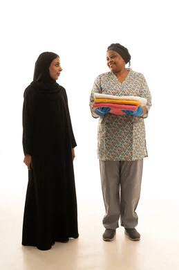 A Saudi Arabian Gulf woman wearing a black abaya and a hijab is observing the work of a housemaid. It is a full-body portrait against a white background of a dark-skinned African maid wearing a gray uniform, holding folded towels.